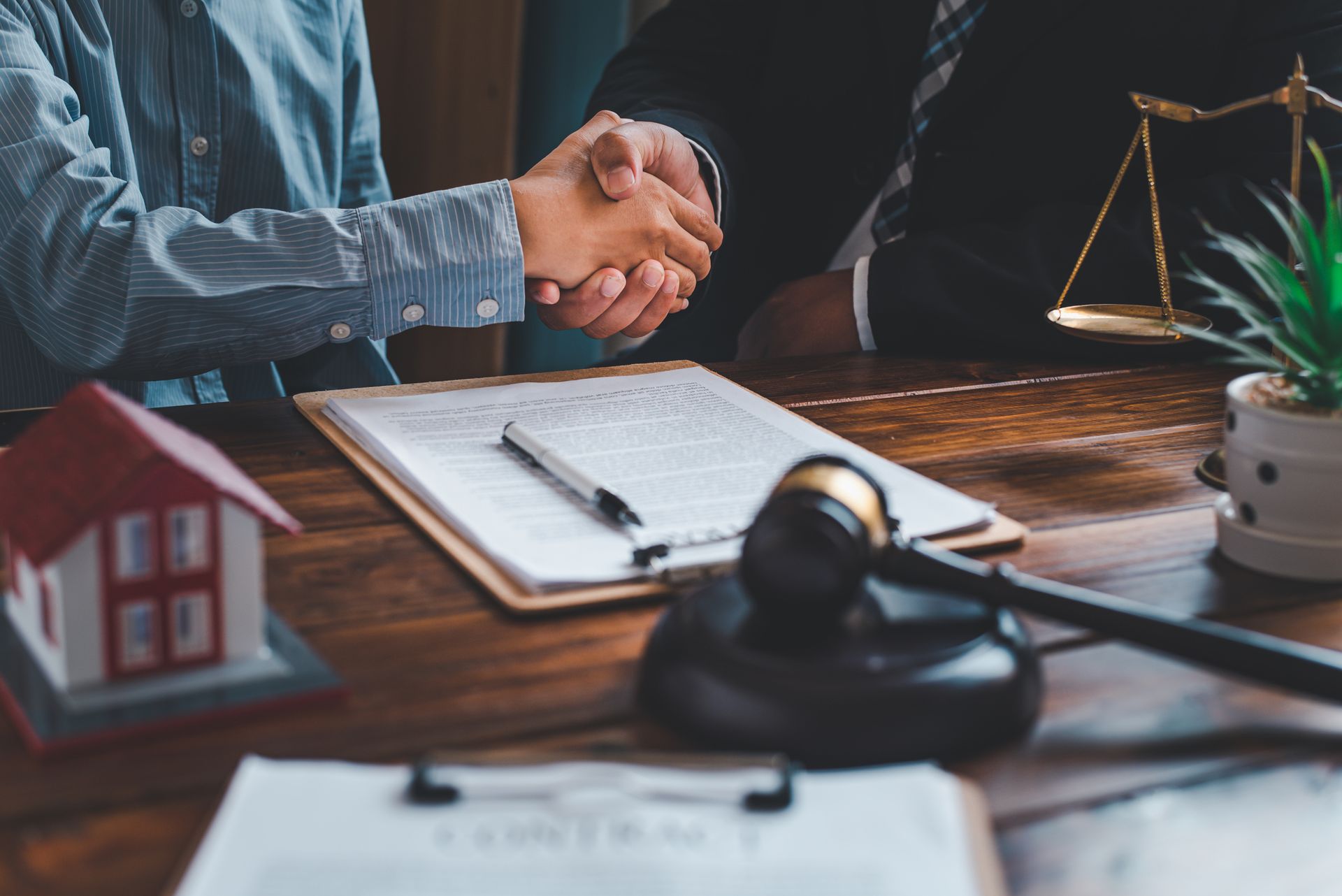Two people shaking hands over a contract, a gavel, and a model house on a desk Two people shaking hands over a contract, a gavel, and a model house on a desk