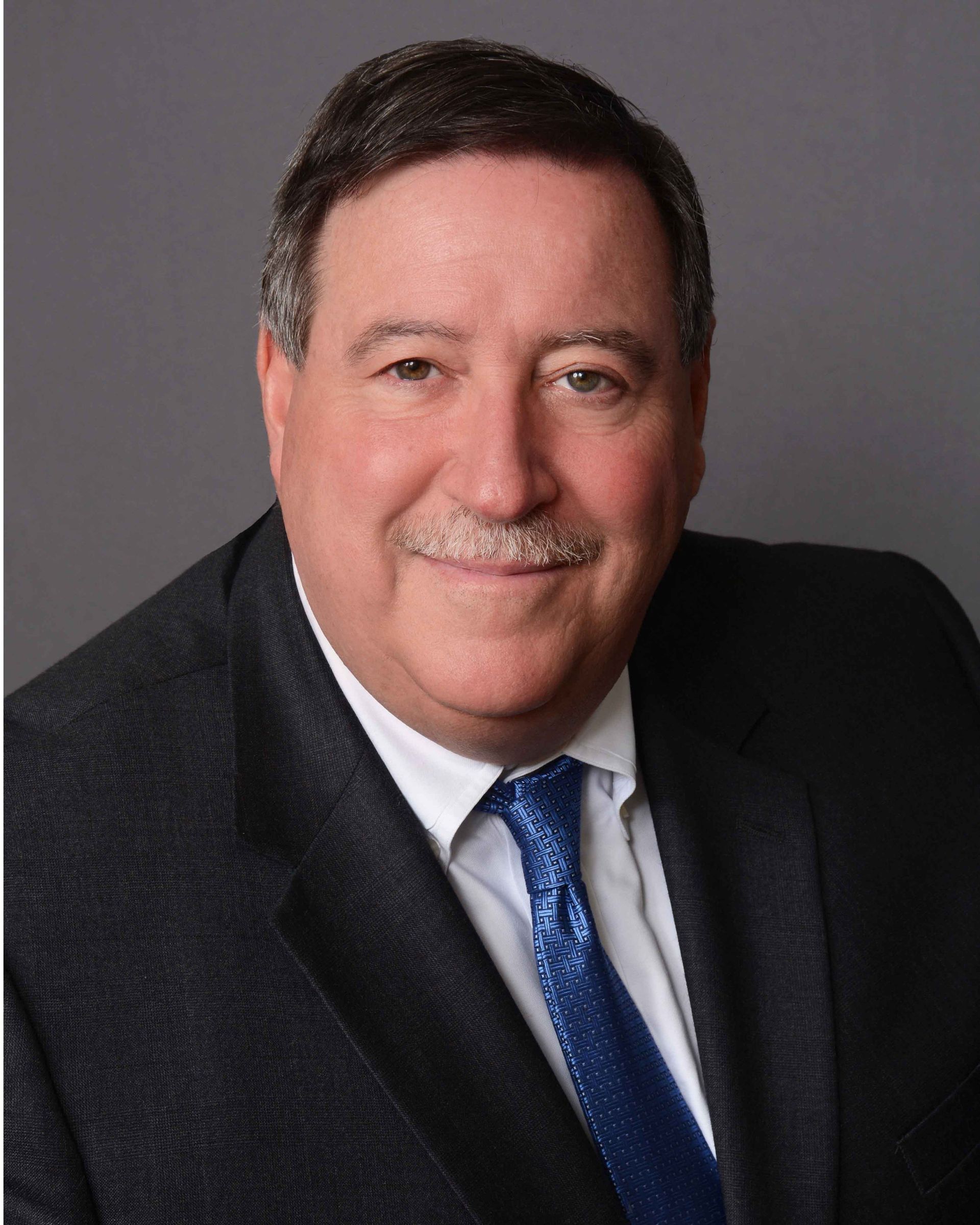 Man in suit and tie with a mustache, smiling, posing for a headshot against a gray backdrop. Man in suit and tie with a mustache, smiling, posing for a headshot against a gray backdrop.