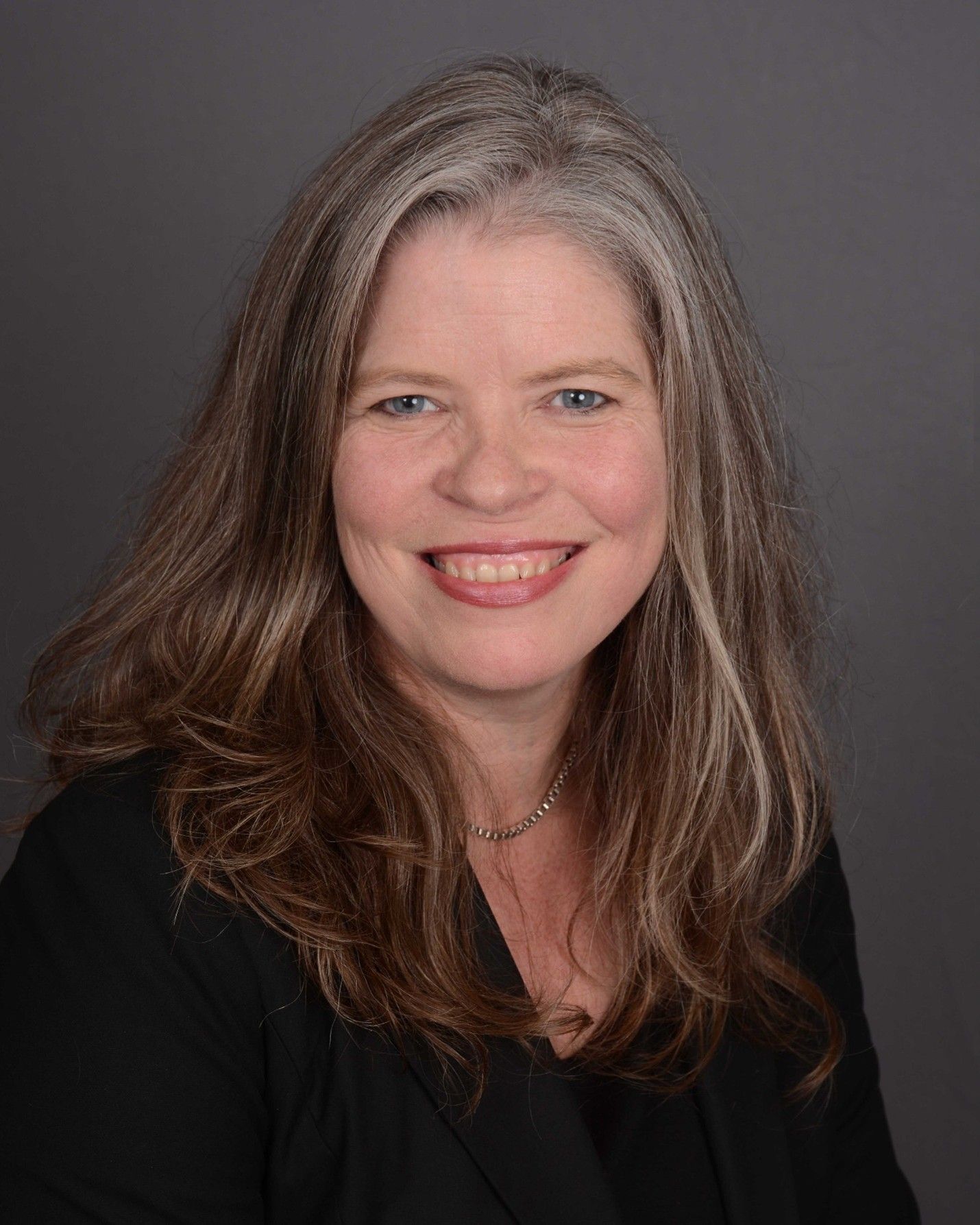 Woman with long graying brown hair, smiling, wearing a black blazer against a gray backdrop. Woman with long graying brown hair, smiling, wearing a black blazer against a gray backdrop.
