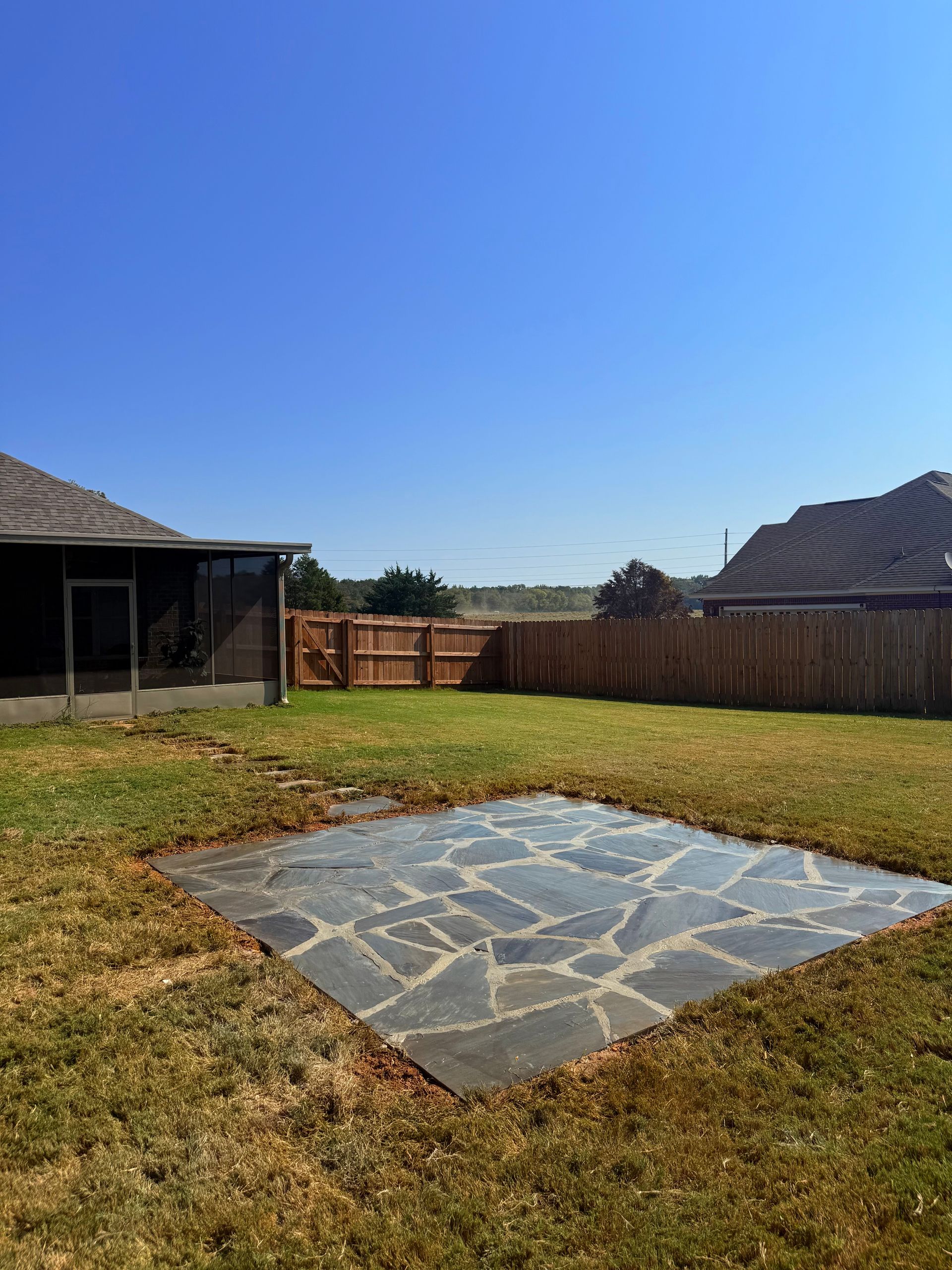 Backyard with flagstone patio, green grass, wooden fence, and clear blue sky.