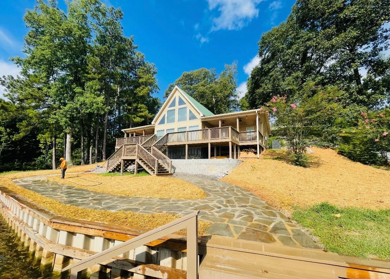 Lakefront house with wooden deck, stone path, and gravel landscaping under a blue sky.