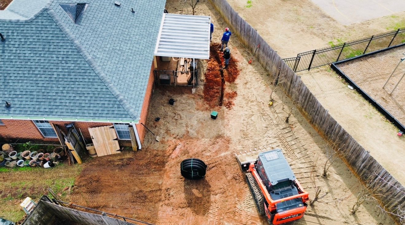 Aerial view of a construction site. A trench is being dug near a house, with an excavator and workers present.