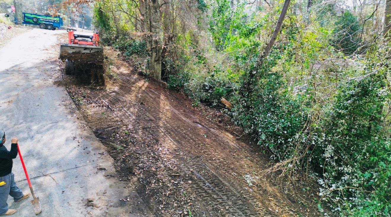 A small tractor spreads mulch along a road, near dense greenery. A worker stands nearby with a shovel.