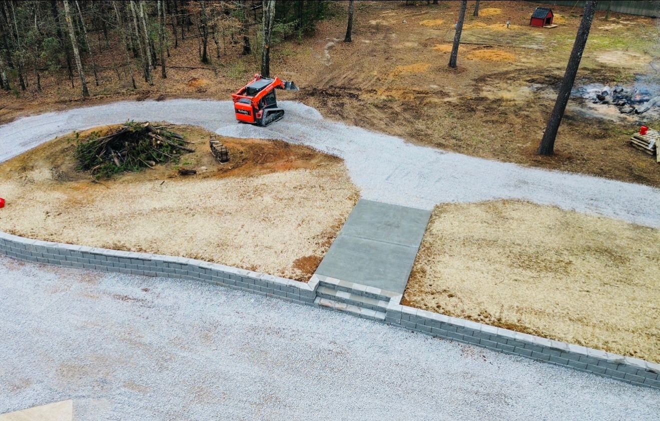 Gravel driveway with retaining walls, small bridge, and an orange skid steer working. Surrounded by wooded area.