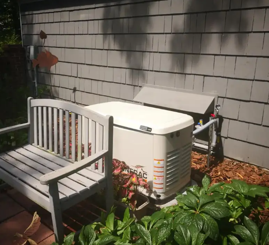 A standby generator next to a wooden bench against a gray house exterior. Plants and wood chips are nearby.
