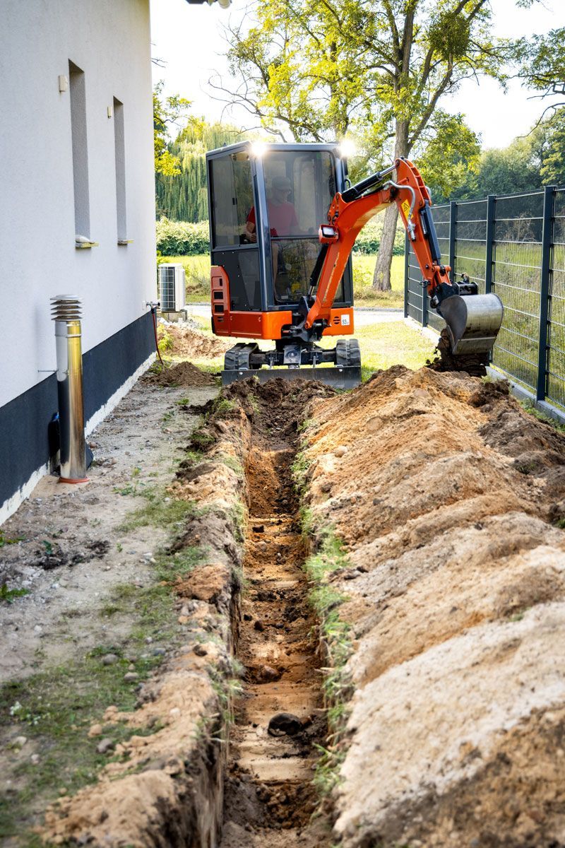 Mini excavator digging a trench beside a building. Orange and black machine, dirt, grass, sunny day.