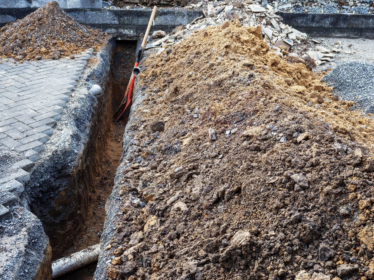 Trench dug in soil, with a shovel resting inside and piles of dirt on the sides near a paved area.