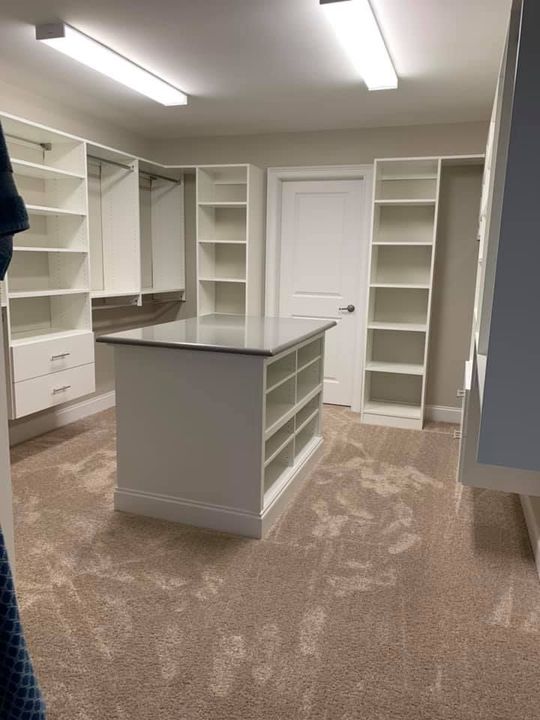 Walk-in closet with white shelving, hanging rods, drawers, and a central island with shelves on a beige carpeted floor.