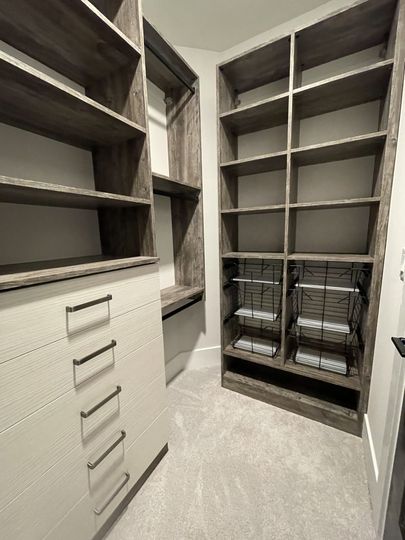 A walk-in closet featuring grey wood shelving, a white six-drawer dresser, and two wire storage baskets on carpet.