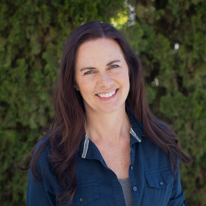 A woman in a blue shirt is smiling in front of a bush.