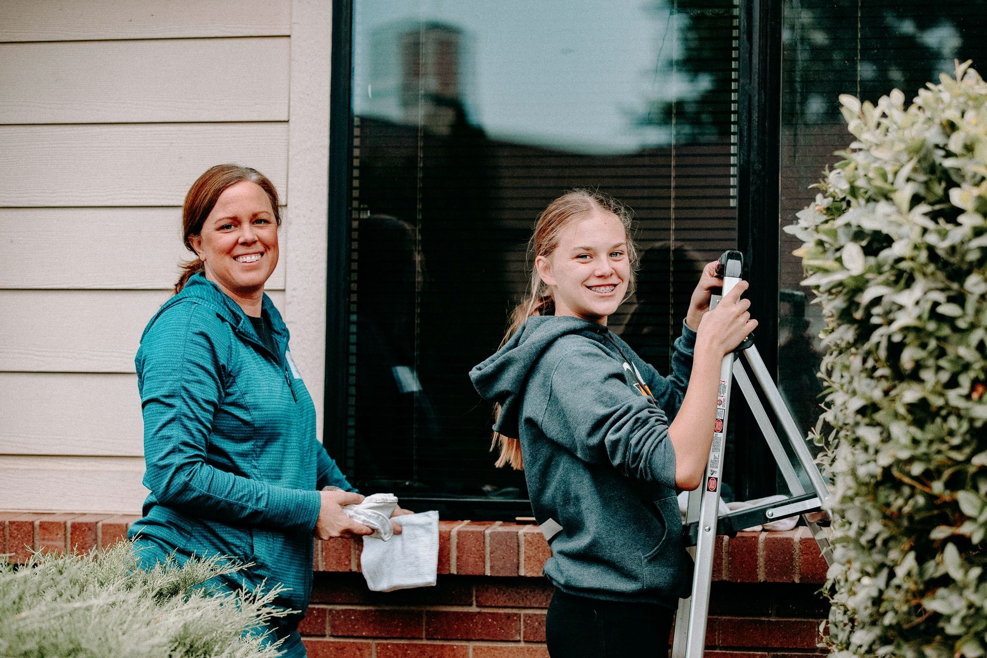 A woman and a teen are standing in front of a window to clean it. 