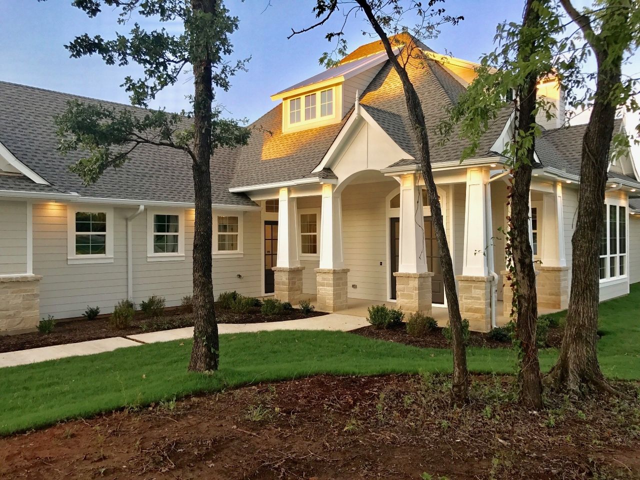 Light-colored house with stone accents, a covered porch, and a lush green lawn. Trees frame the view.