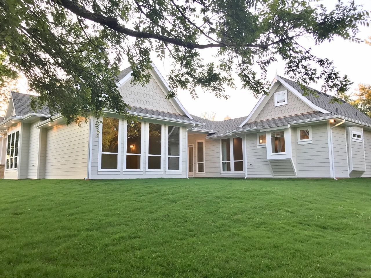 Light-colored house with multiple windows and gabled roof, set on a green lawn beneath tree branches.