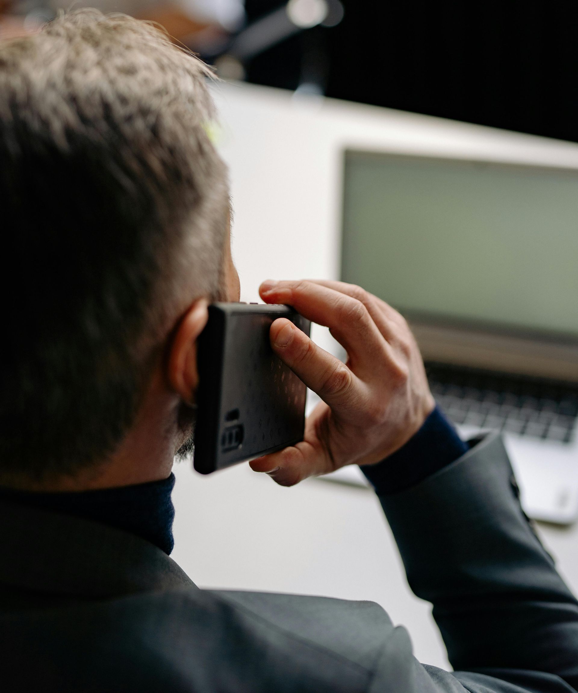 Man on a phone call, holding a dark phone to his ear, near a laptop on a white desk.