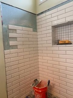Small tiled shower stall with a red bucket and cleaning tools on the floor.