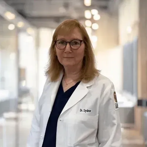 A healthcare professional in a white coat and glasses standing in a professional clinic or office hallway.