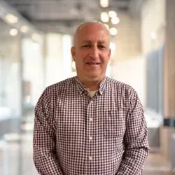 A person wearing a maroon and white checkered button-down shirt, standing in a blurred indoor office hallway.