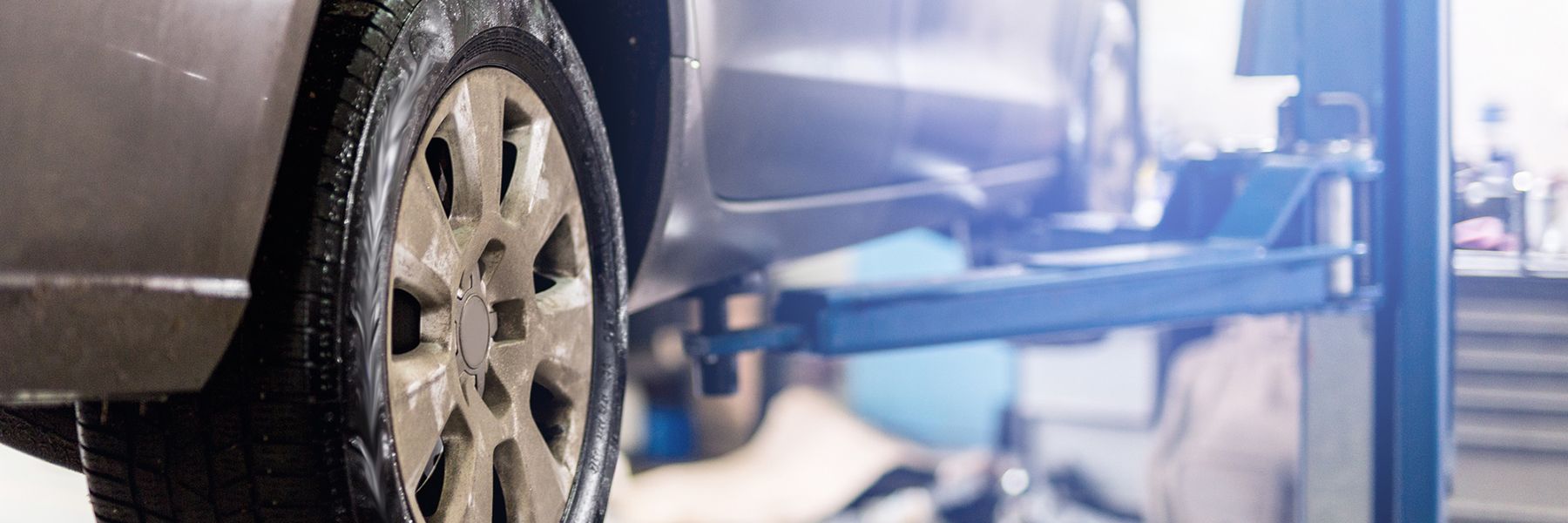 Car tire raised on a lift in a garage. Close-up of tire with dirty rim. Mechanics working on the car.