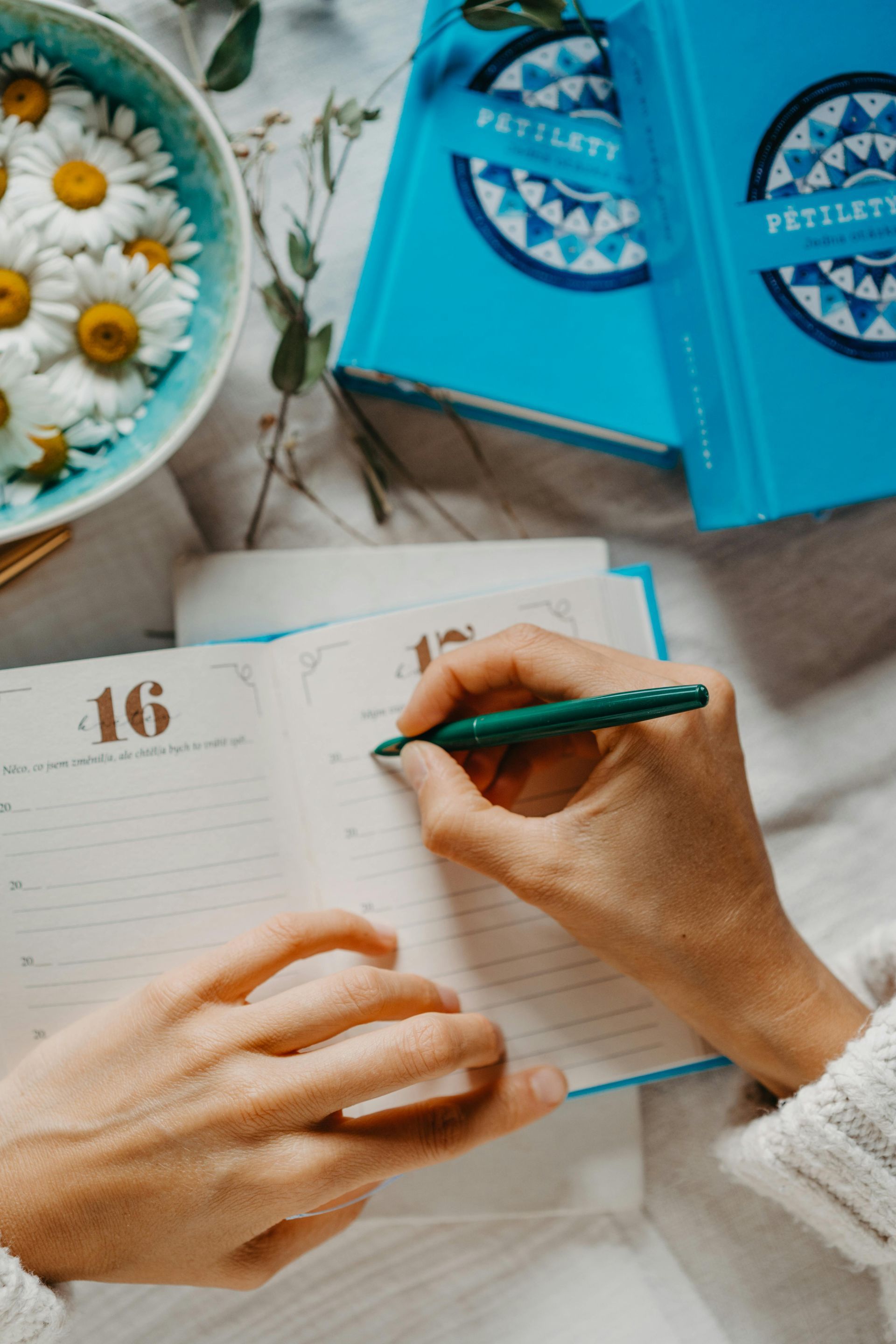 Uma pessoa escreve em uma agenda datada do dia 16, com uma tigela de margaridas e dois cadernos azuis sobre uma superfície branca próximos.