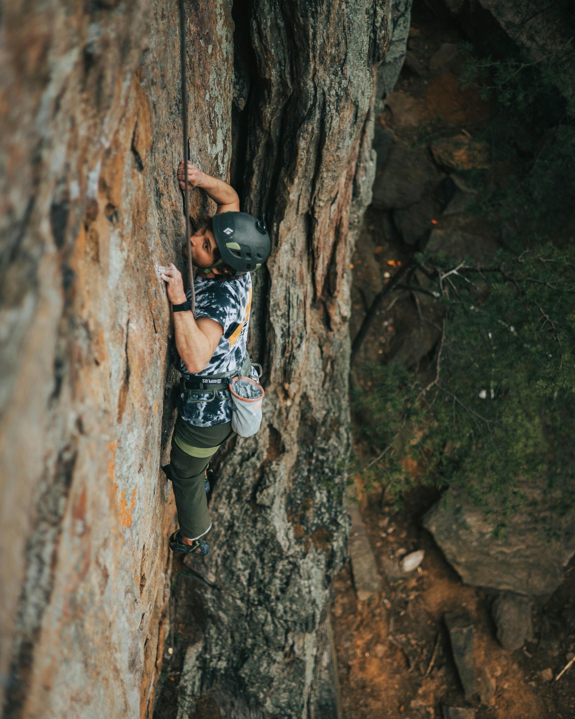 Um alpinista usando capacete e arnês escala uma parede rochosa íngreme e vertical, com as mãos buscando um ponto de apoio.