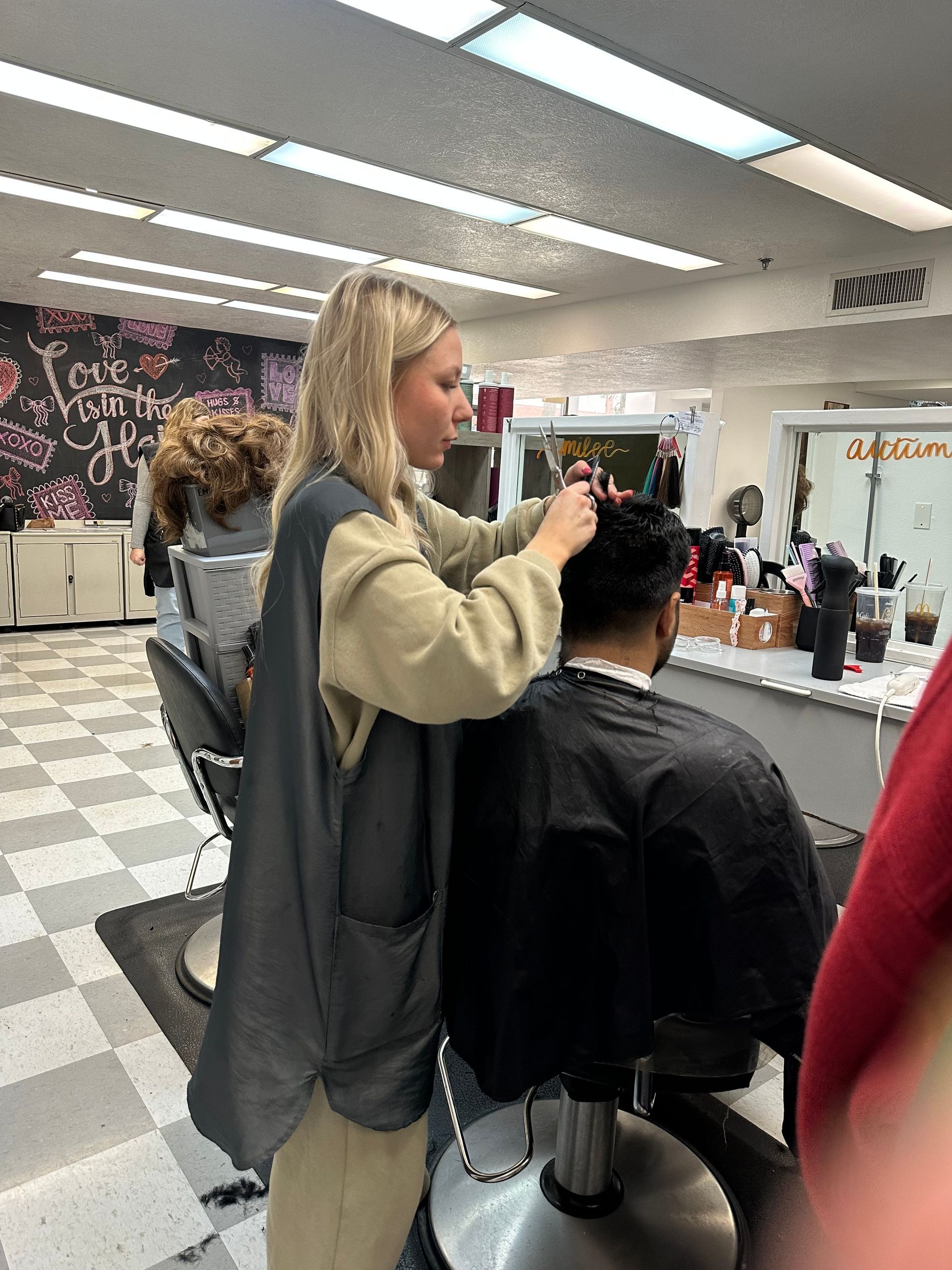 A stylist in a tan top and gray apron cuts the hair of a person wearing a black cape inside a salon.