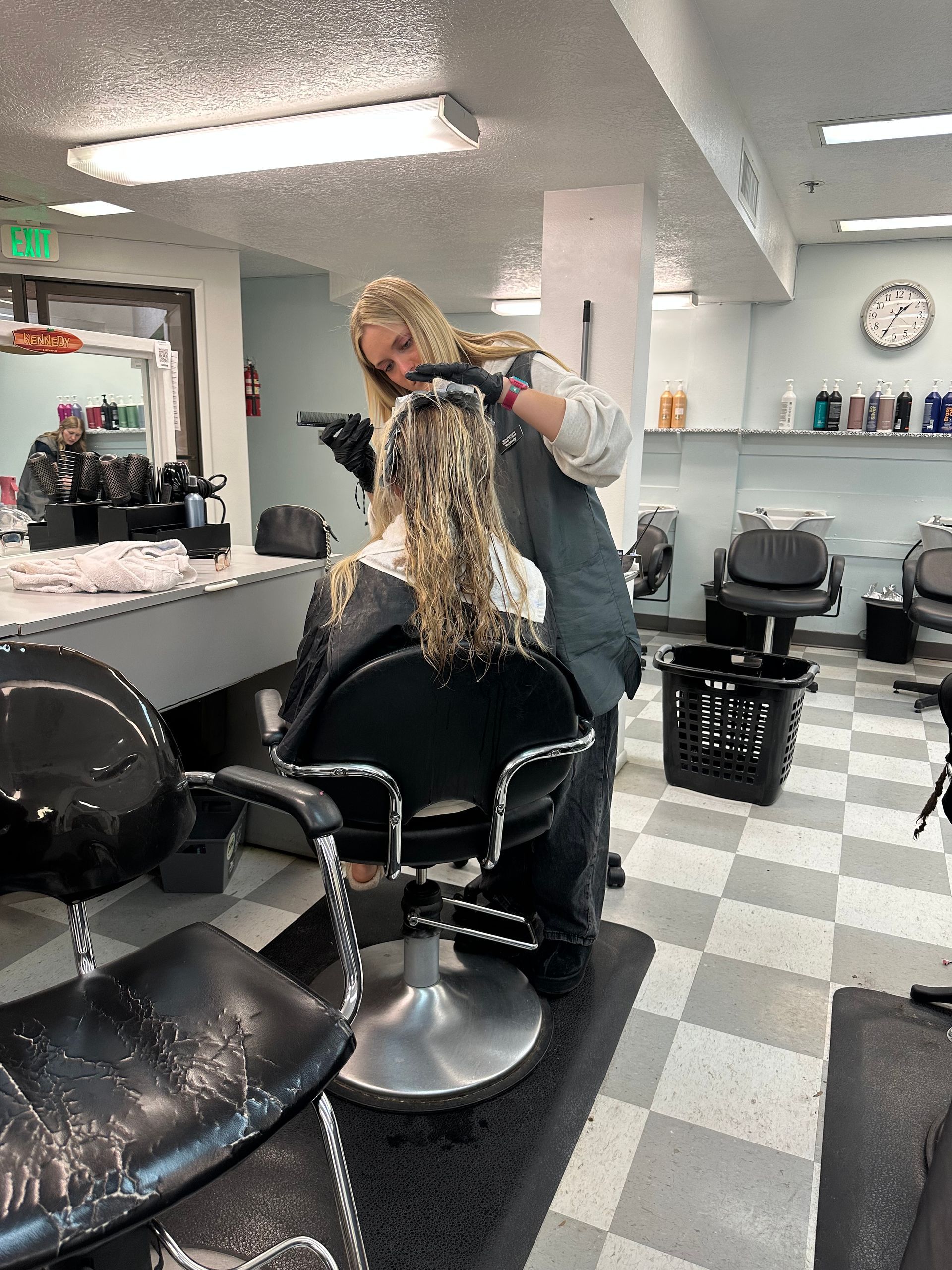 A hair stylist applies treatment to a client’s curly hair in a salon with tiled floors and chairs.