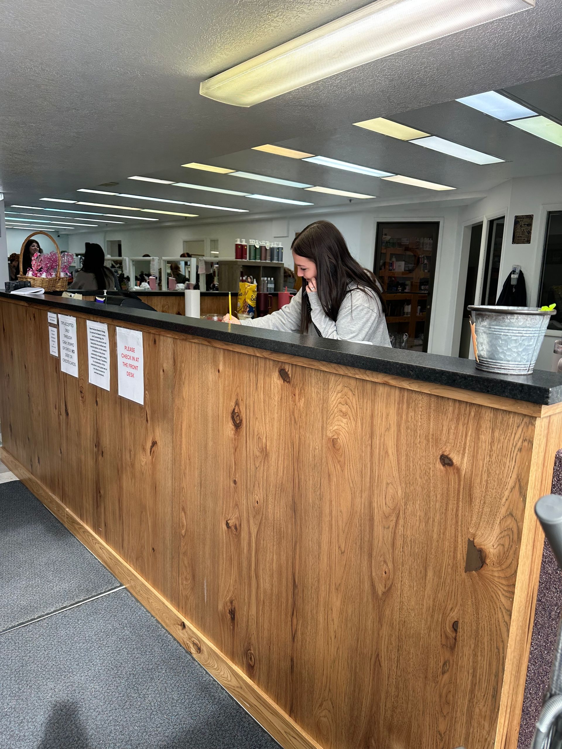 A person works behind a wooden reception desk in a brightly lit, commercial office or salon space.