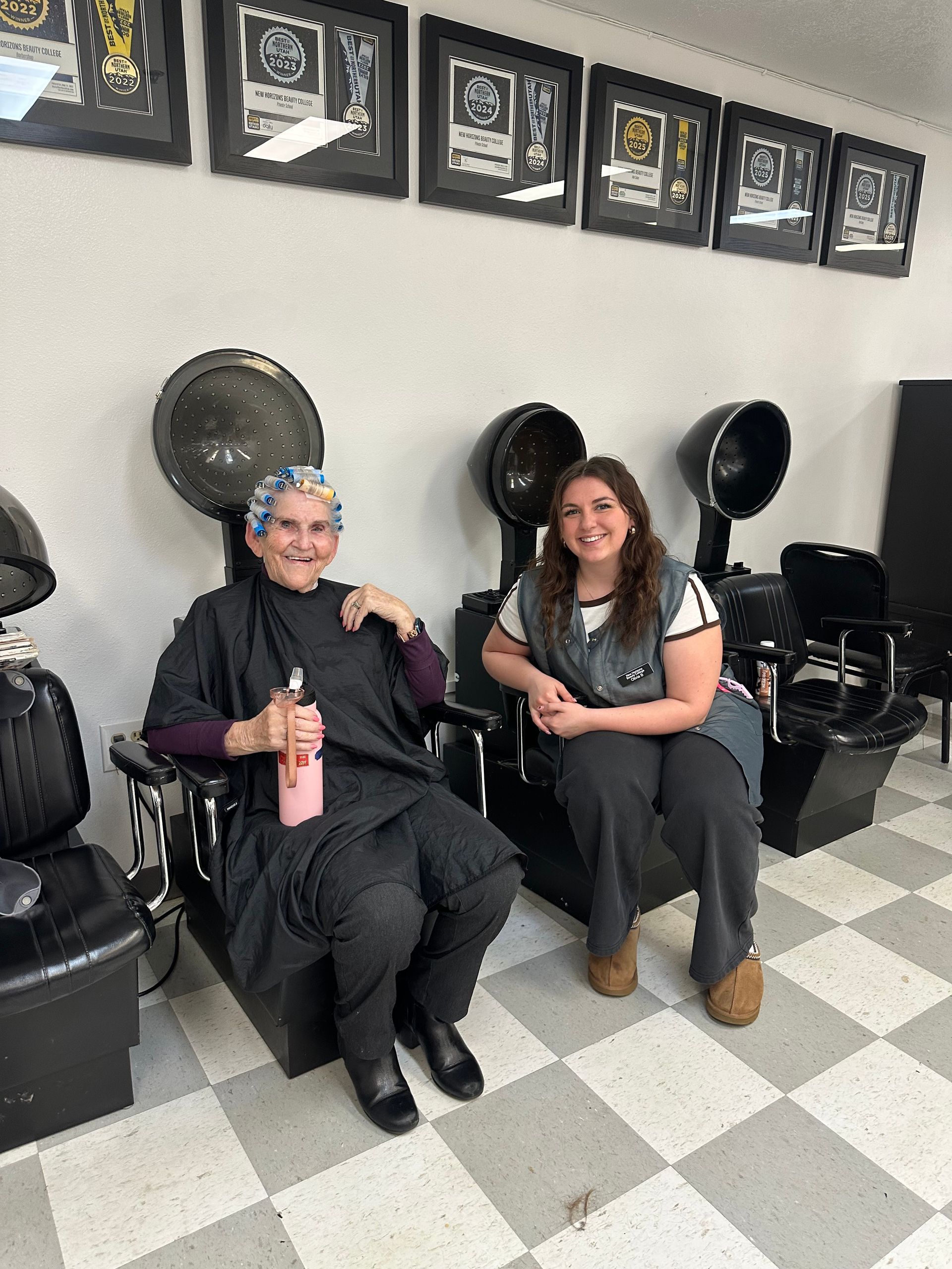 Two people sit in a salon under hooded hair dryers, smiling in front of a wall decorated with framed certificates.