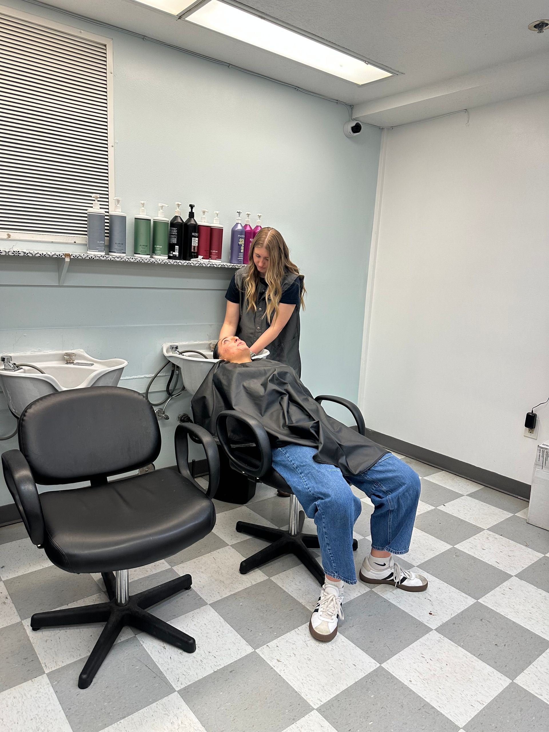 A stylist attends to a client having their hair washed at a salon sink, both seated in chairs in a checkered-floor room.
