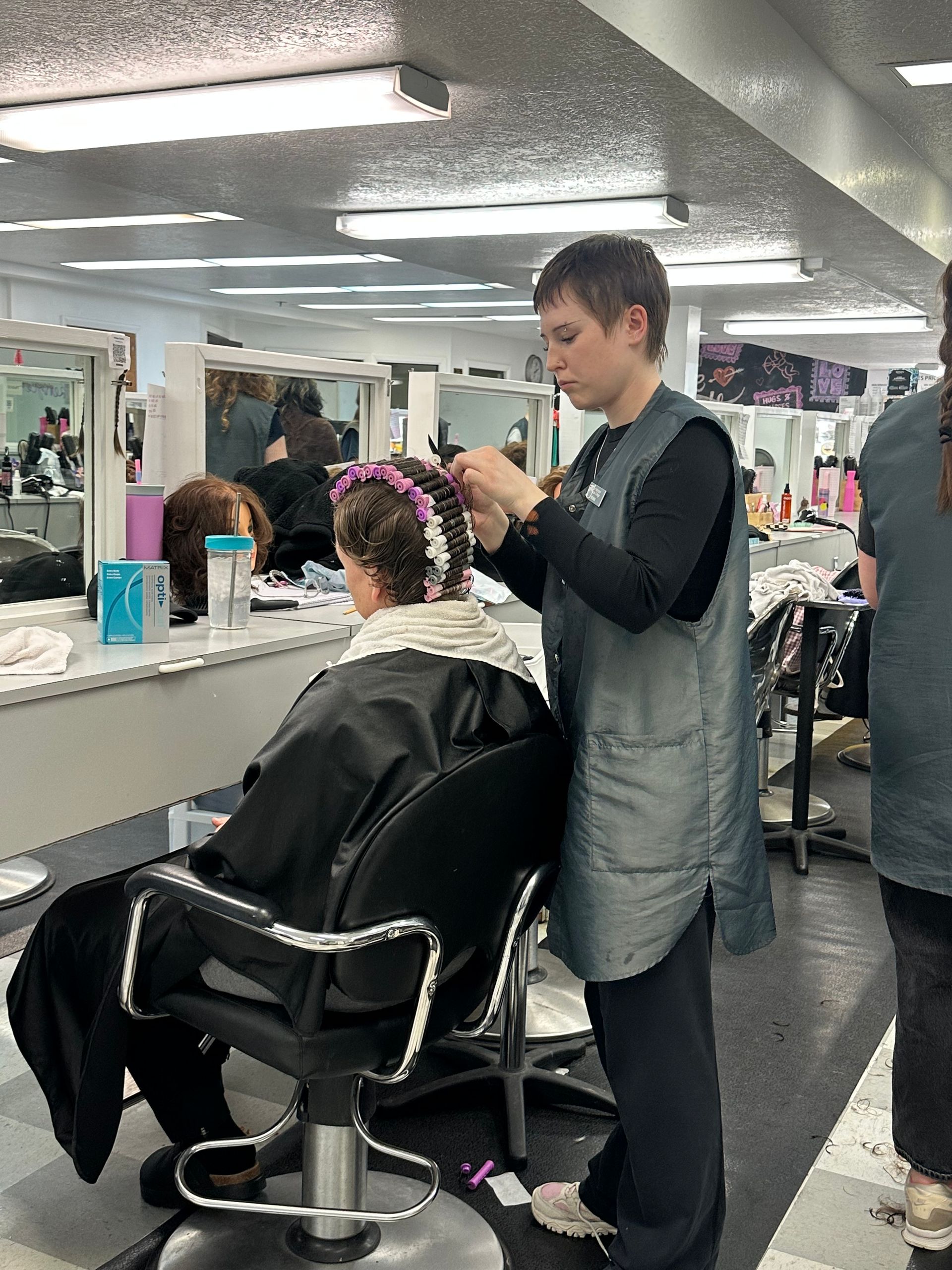 A student stylist works on a client’s hair in a cosmetology classroom with multiple workstations and mirrors. A student stylist works on a client’s hair in a cosmetology classroom with multiple workstations and mirrors.