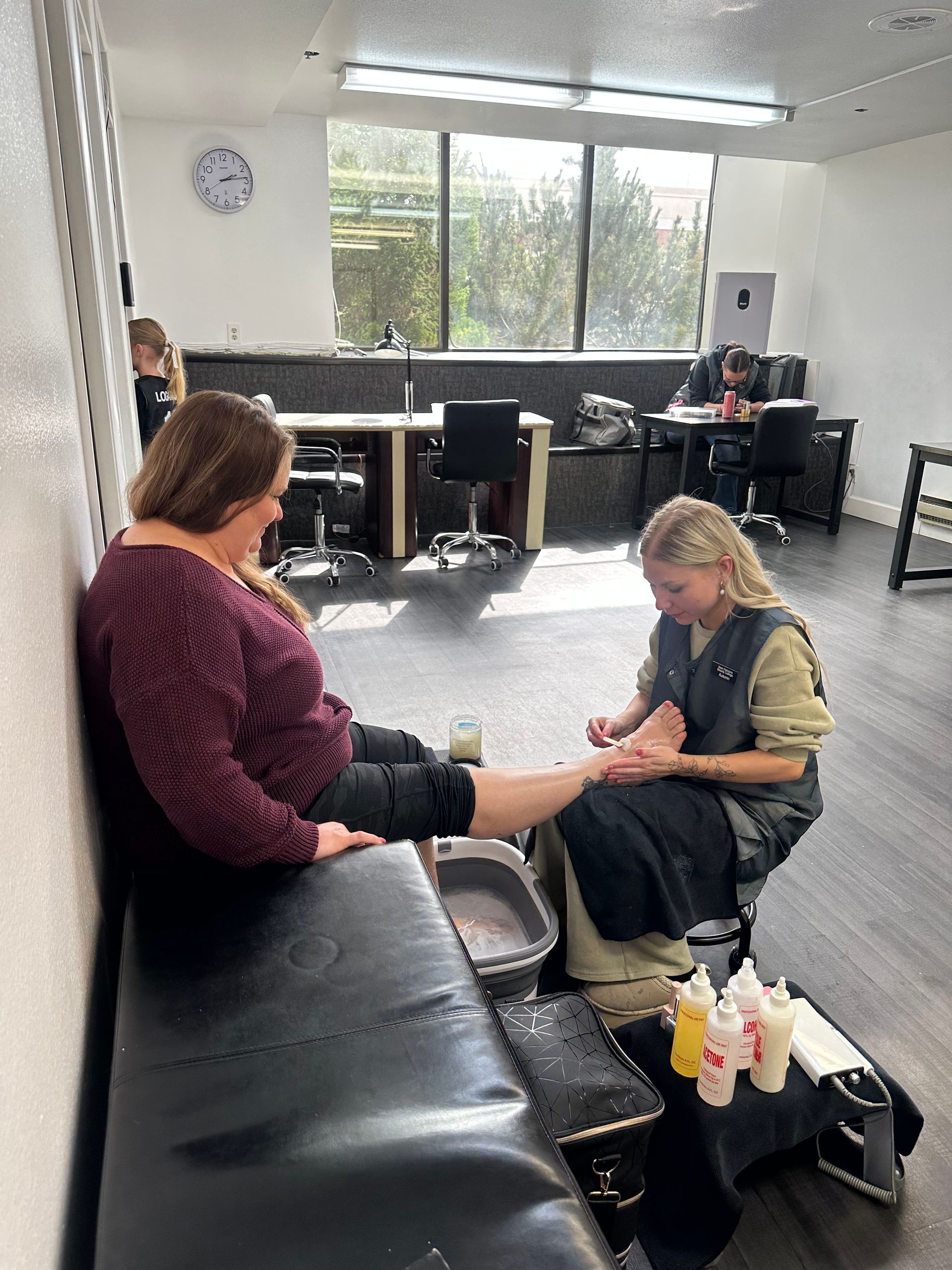 A professional performs a pedicure on a client sitting on a black bench in a well-lit salon with workstations. A professional performs a pedicure on a client sitting on a black bench in a well-lit salon with workstations.