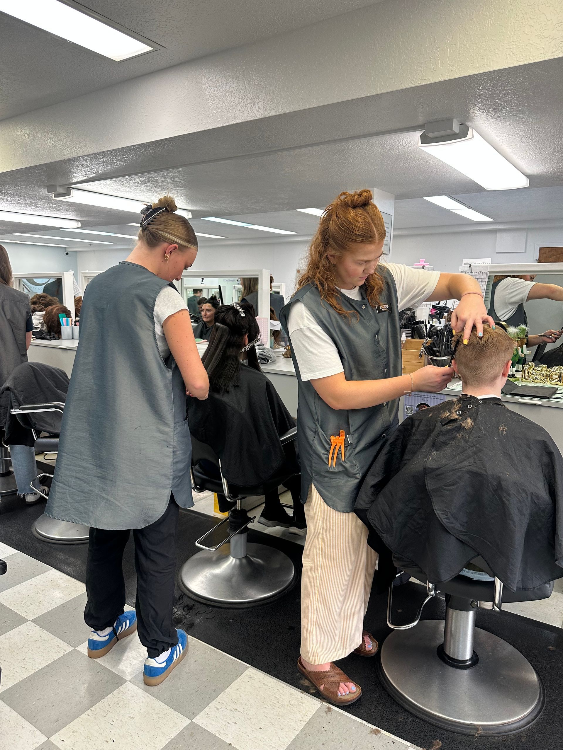Two students in gray salon vests cut hair in a bright, tiled classroom setting.