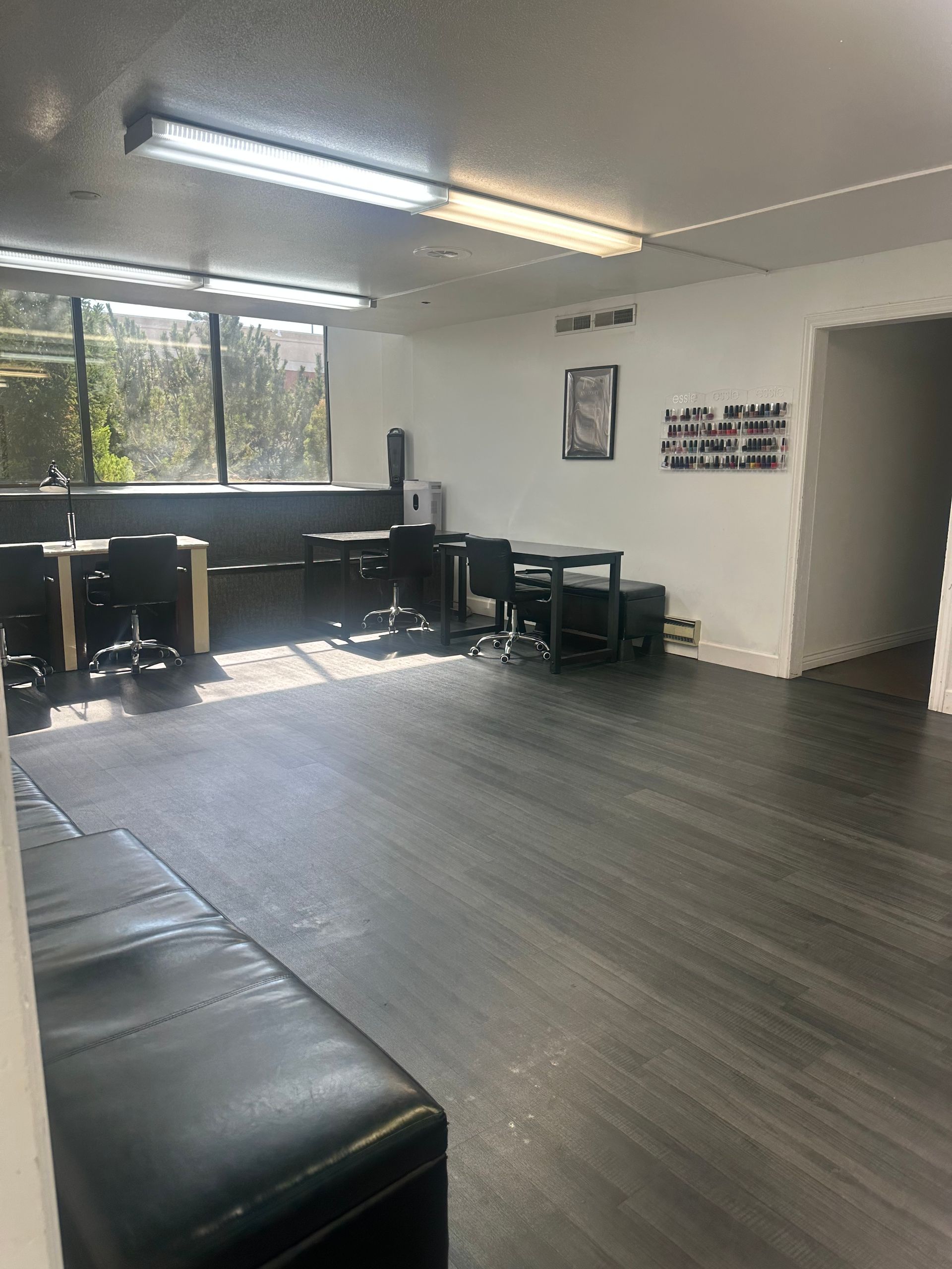 An empty, brightly lit office or salon space with wood-style flooring, black chairs, tables, and a bench along the wall.
