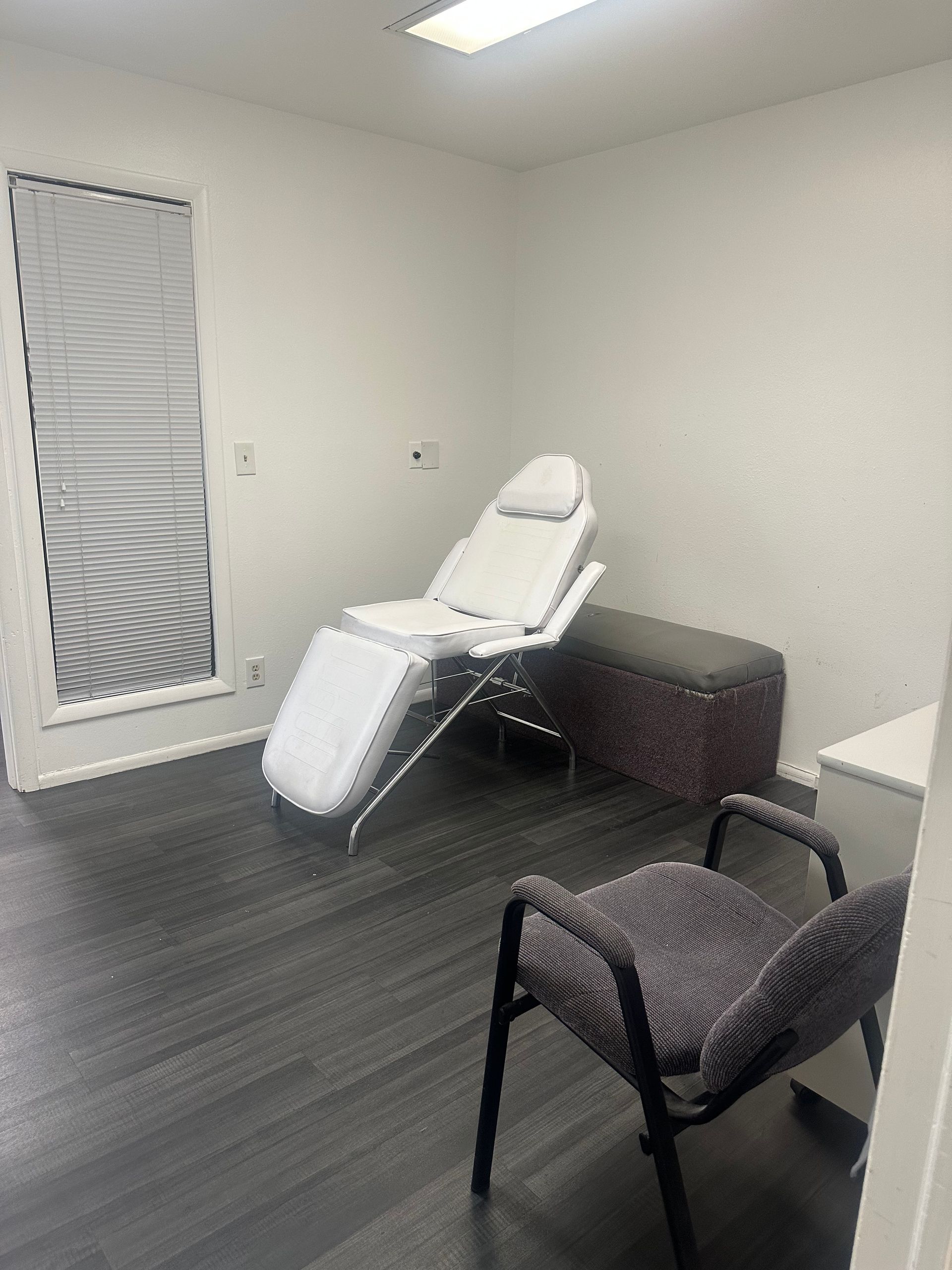 A medical treatment room featuring a white reclining exam chair, a grey upholstered bench, and a patterned task chair.