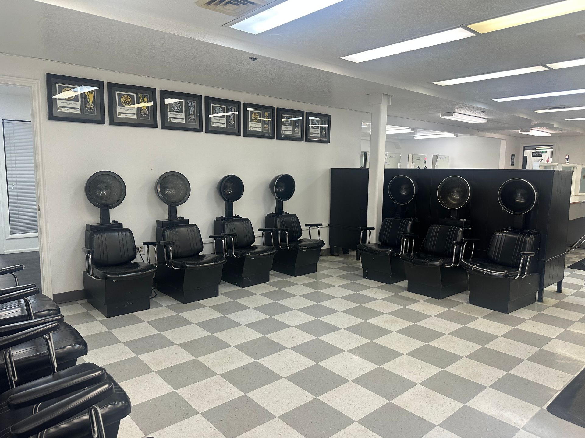 A hair salon interior with two rows of black dryer chairs facing opposite directions on a gray and white checkered floor.