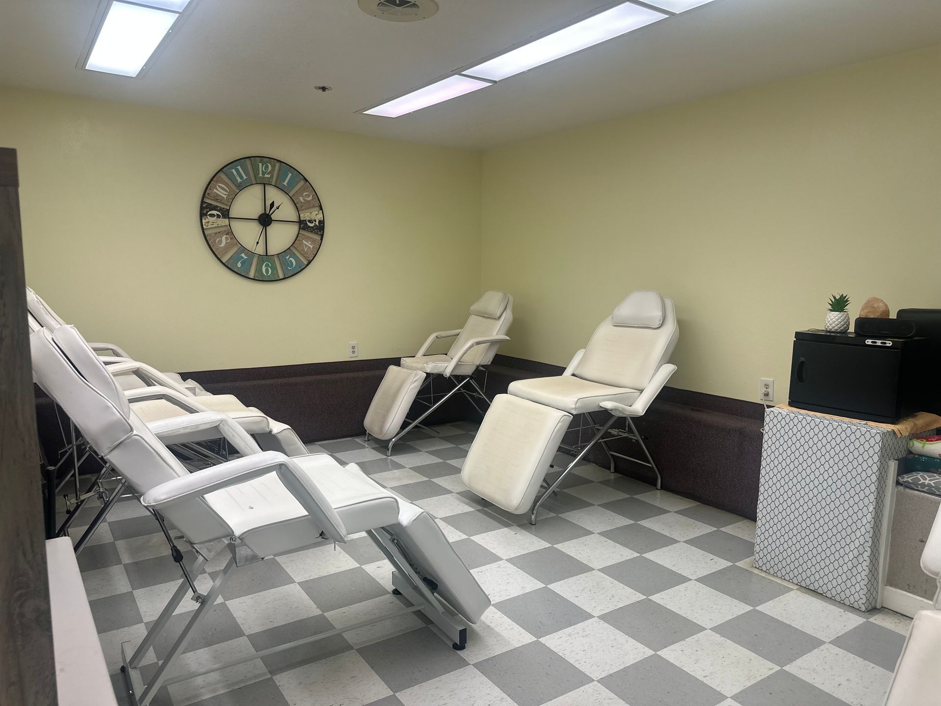 A room with light-colored walls and a checkered floor, featuring four adjustable spa chairs and a large wall clock.