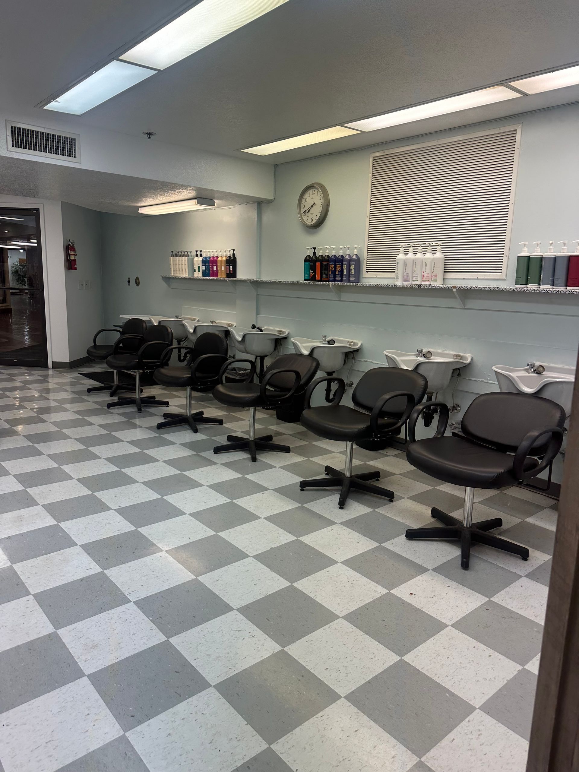 A row of black salon chairs and shampoo sinks in a bright room with a checkered floor and a large abstract wall art.