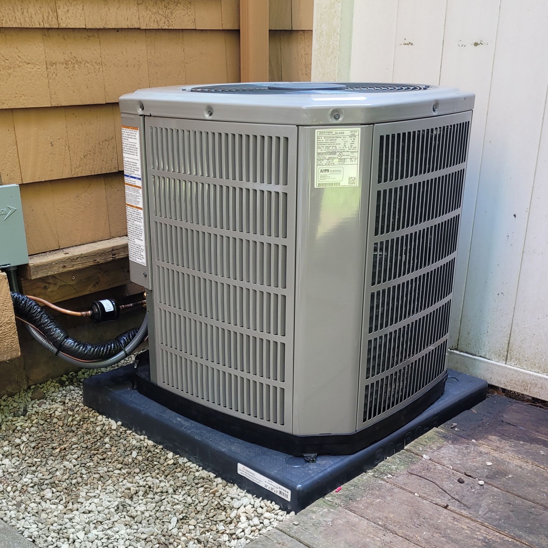 Gray air conditioning unit on a black pad, set on gravel and wooden deck near a wall.