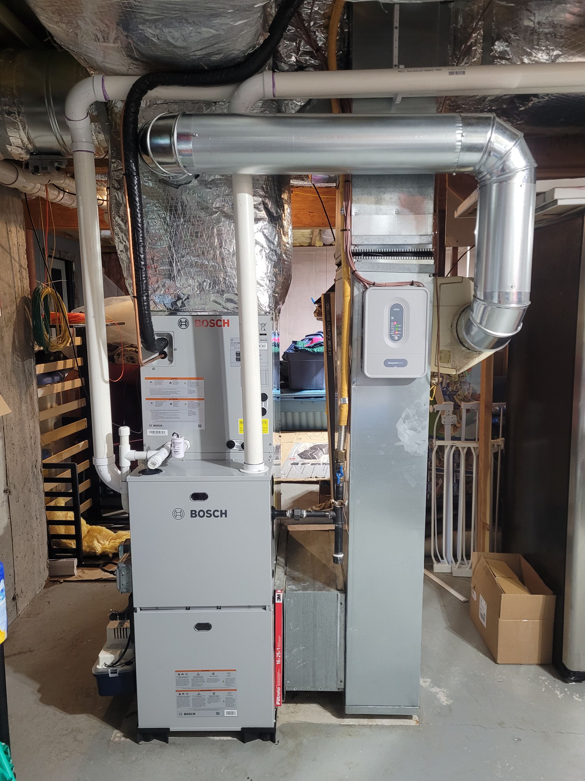 HVAC system: gray furnace with ductwork against a wall. Silver ventilation, white pipes, and cardboard box.