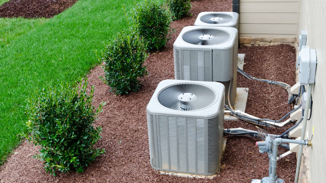 Air conditioning units lined up next to a building on a bed of brown mulch, with green bushes and grass nearby.