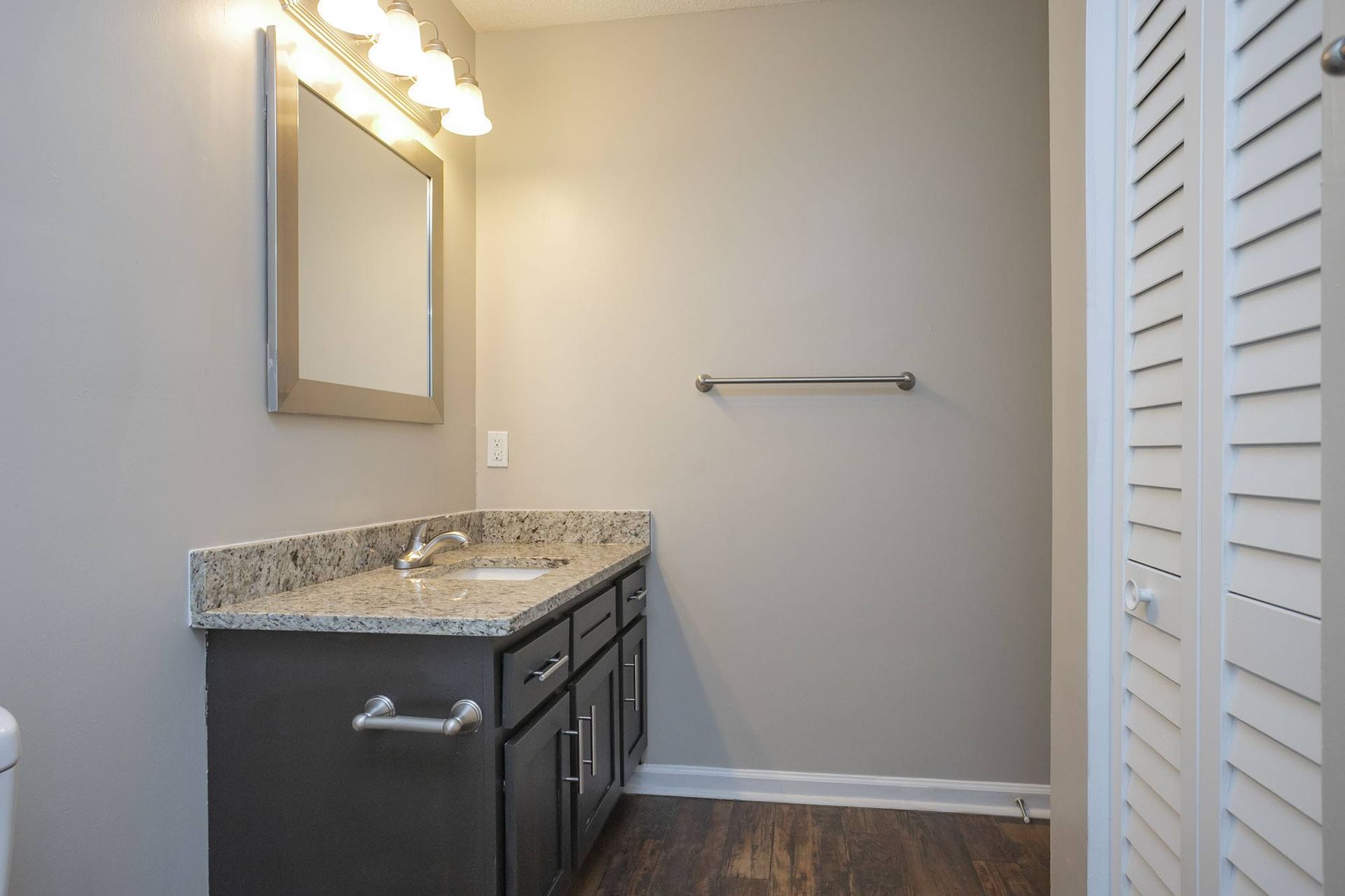 A bathroom vanity with a granite countertop, sink, dark cabinets, a mirror, and light fixtures against a gray wall.