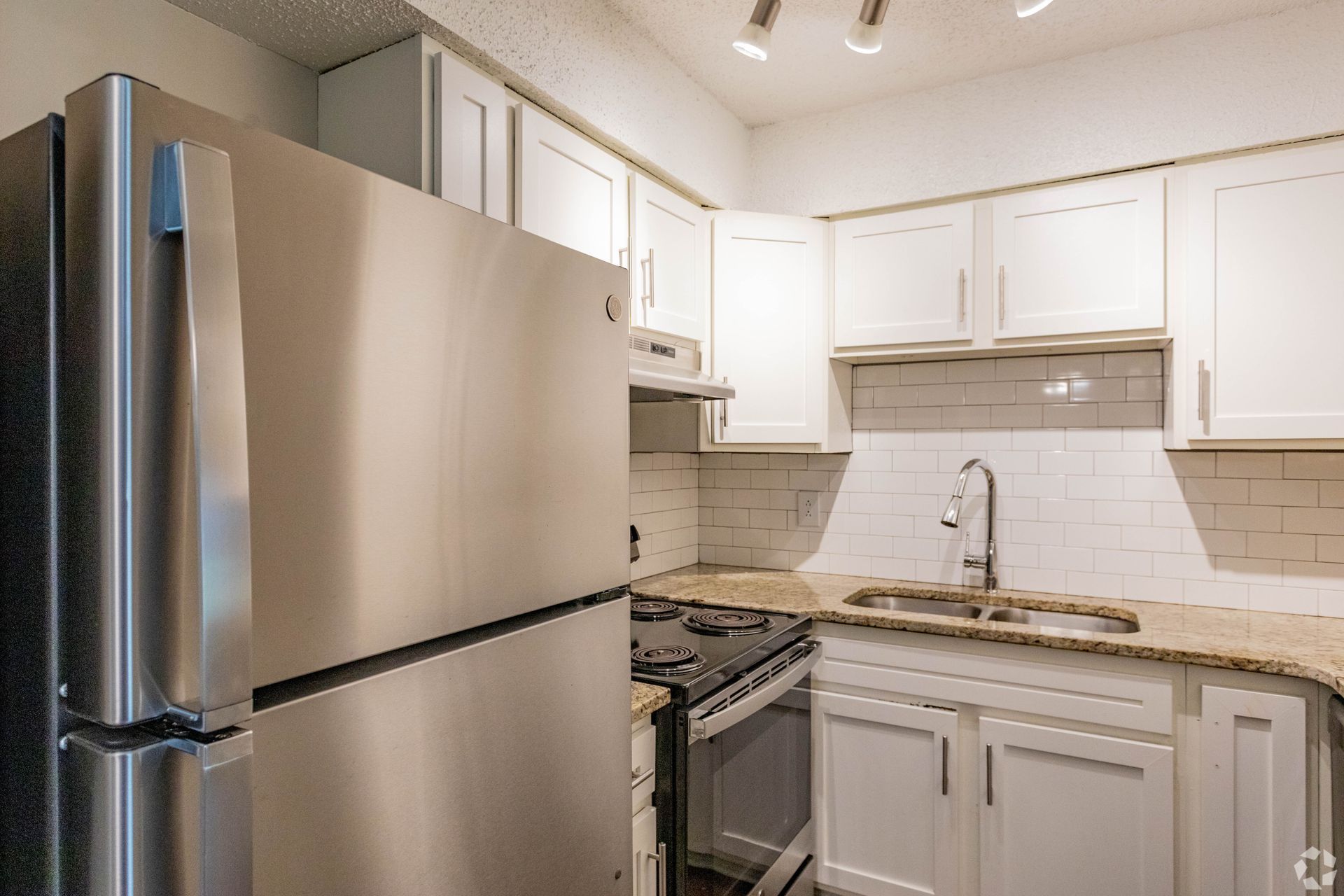 A small, modern kitchen with white cabinets, speckled granite countertops, stainless steel appliances, and tile backsplash.