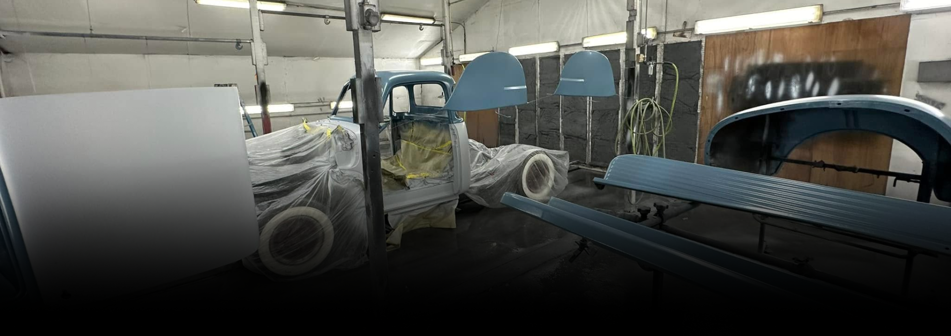A classic blue pickup truck in a body shop, covered with plastic, undergoing restoration.
