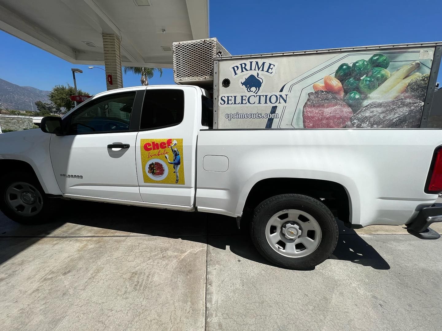 White pickup truck with fruit stand graphics parked at a gas station pump under a canopy.