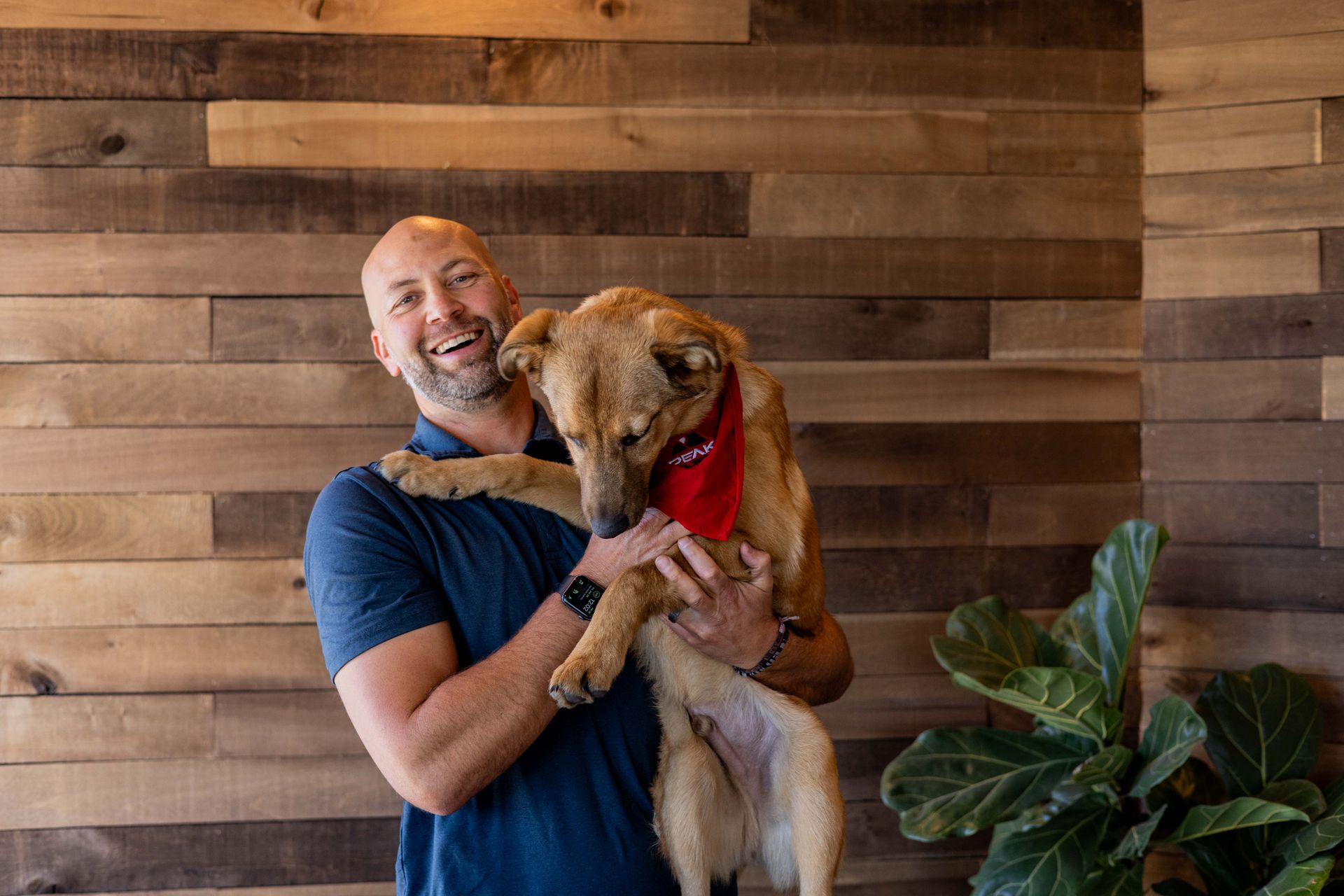 Man holding a dog with a red bandana; both smiling in front of a wood panel wall. | Peak Euro