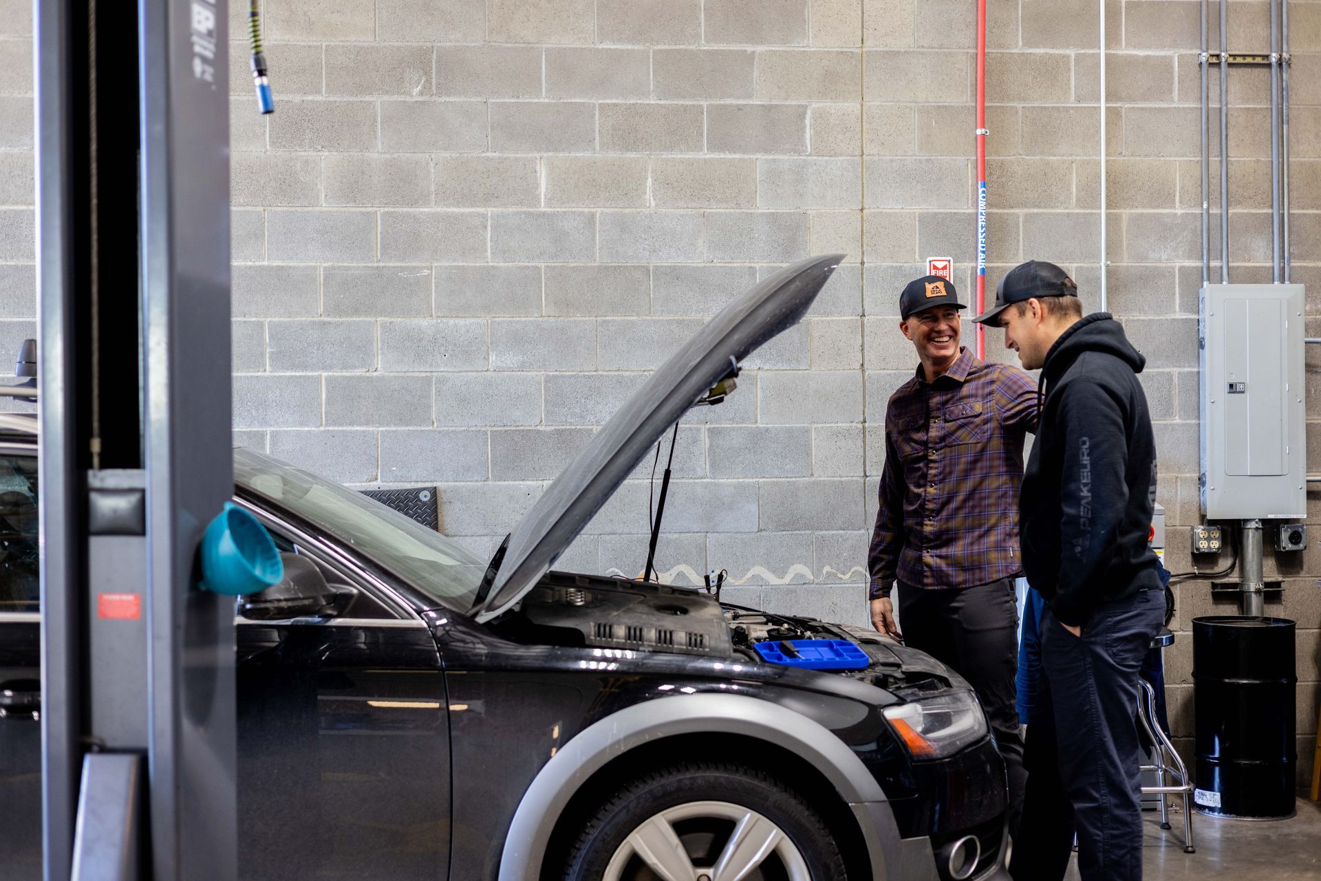 Two men examining a car engine in a garage; one points while the other looks on.
