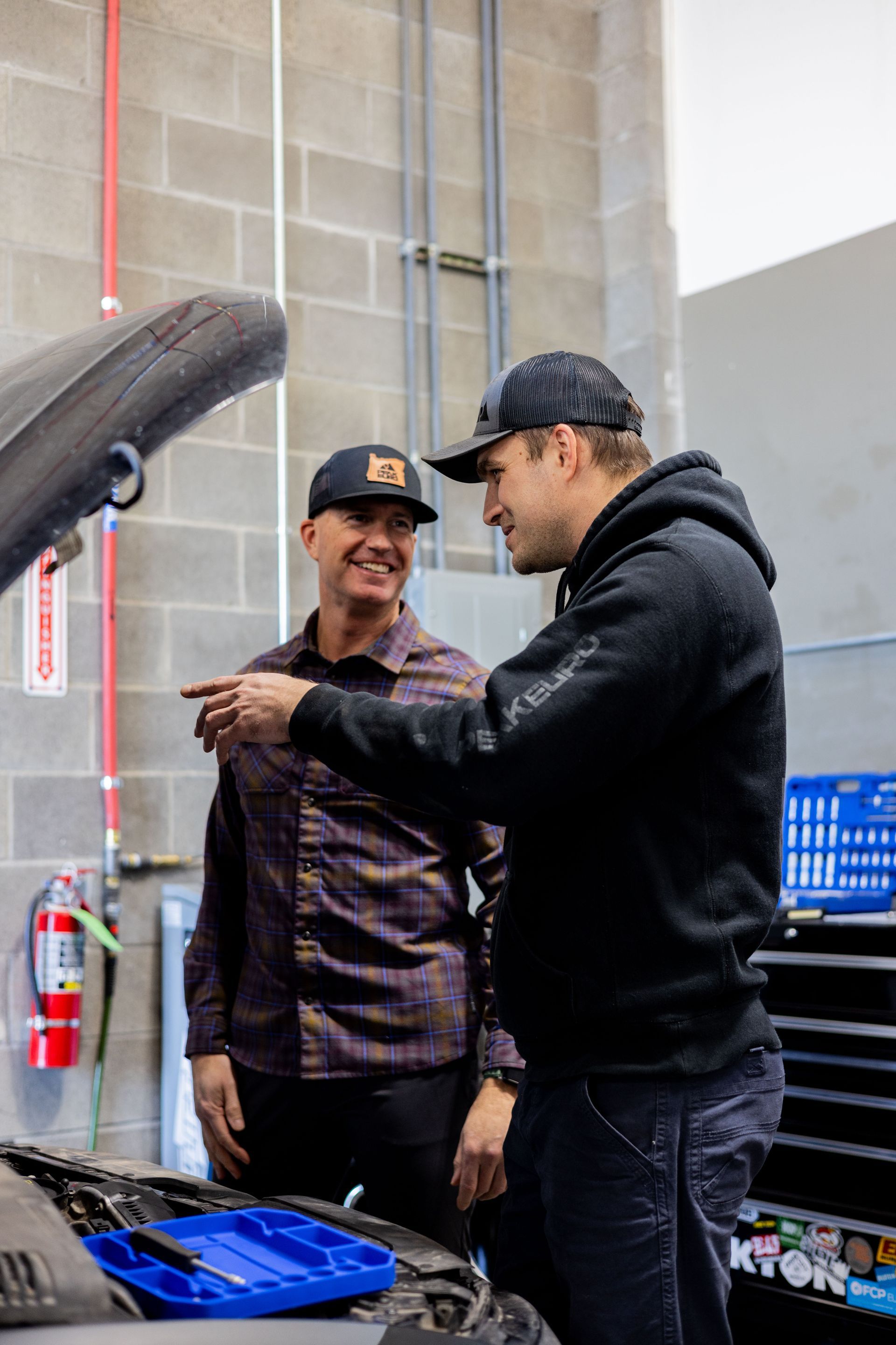 Two men in a garage look at a car engine. One points and explains while the other smiles.| Peak Euro