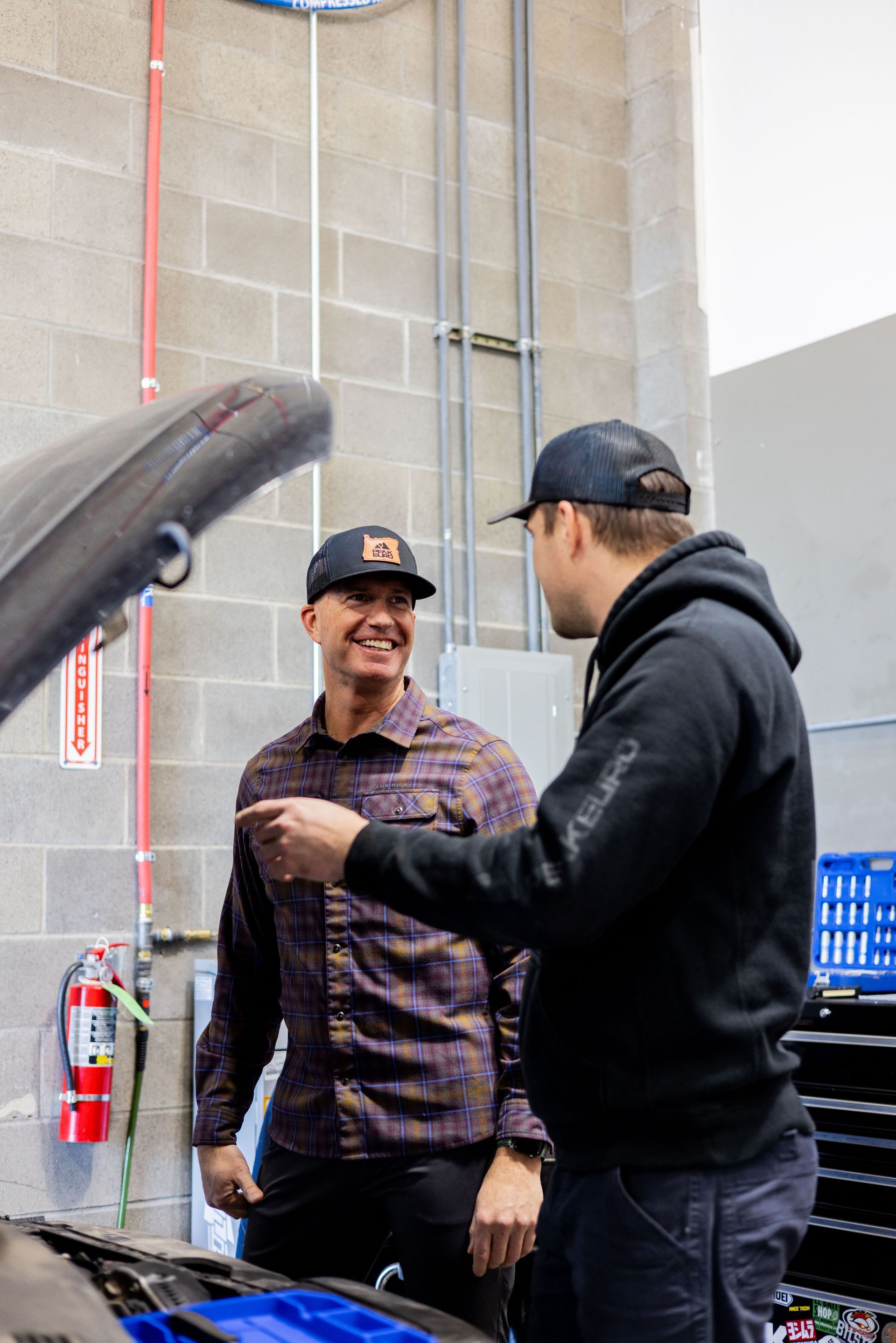 Two men in hats, one pointing near an open car hood, in a garage setting.| Peak Euro