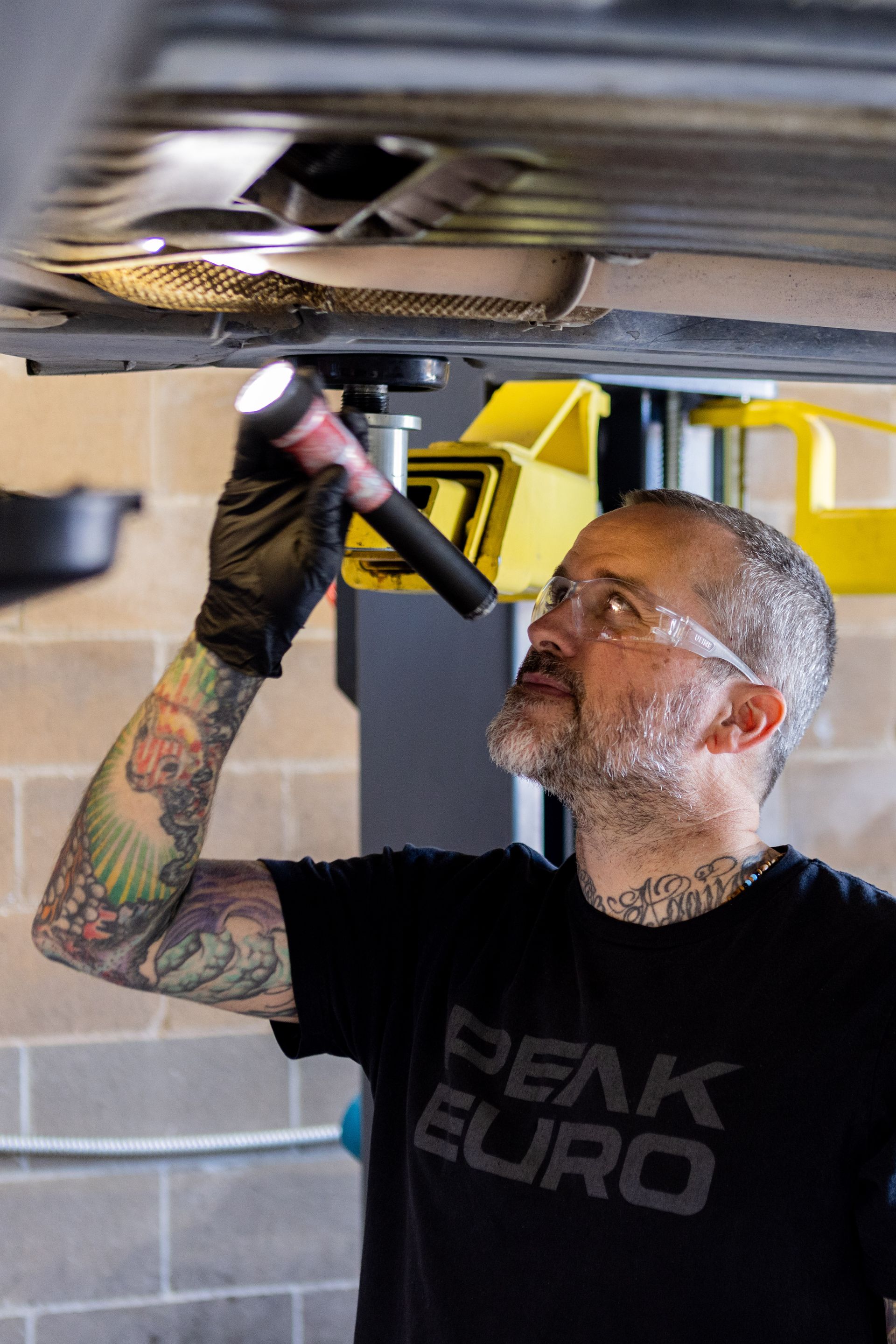 Mechanic inspecting car underside with flashlight, wearing safety glasses and gloves. Garage setting. | Peak Euro