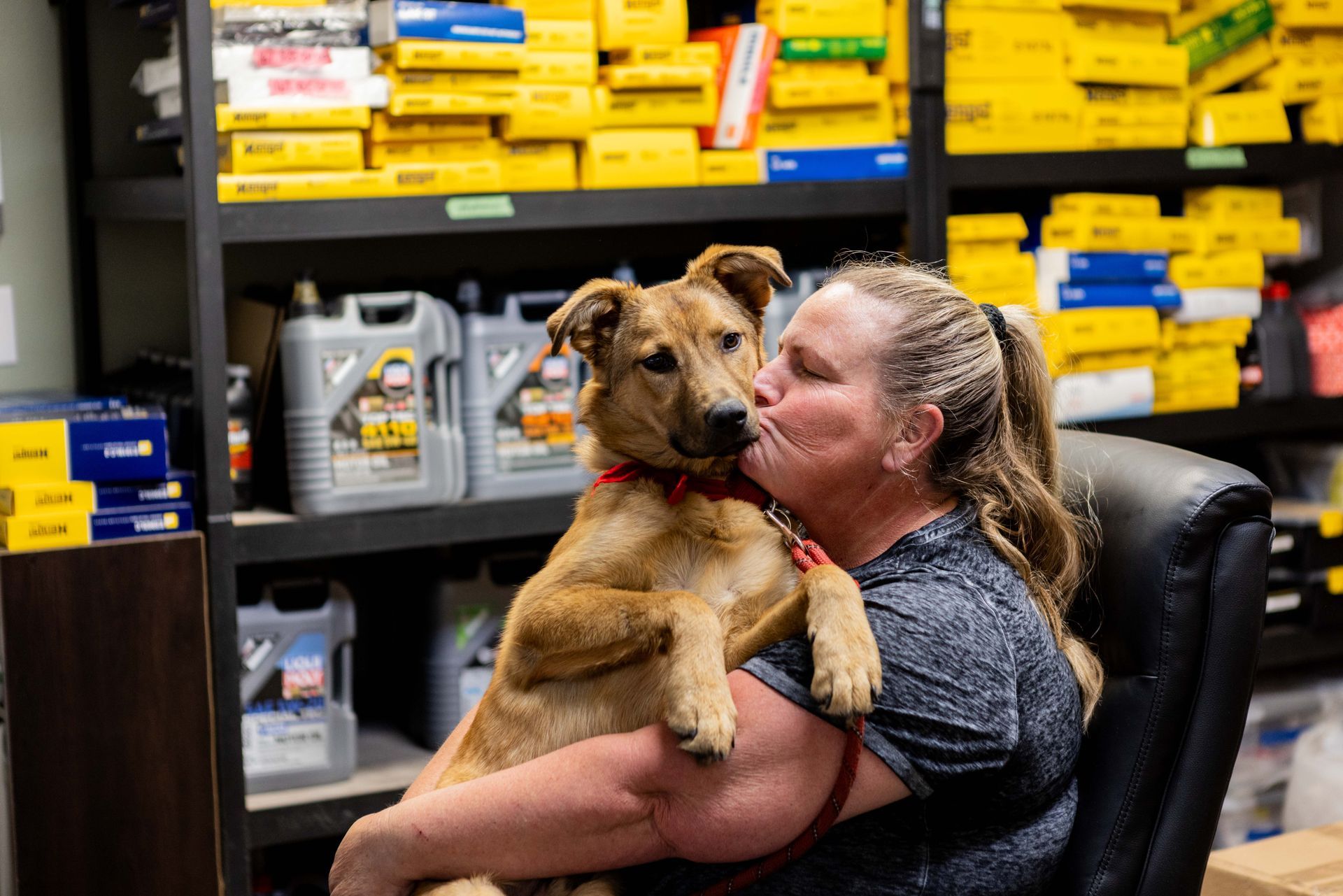 Woman kissing a brown dog. They are in an auto parts store, surrounded by supplies. | Peak Euro